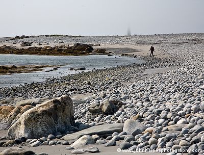 Phyllis hikes along the rocky shore of Cape Sable Island with the lighthouse in the background partially obscured by fog
