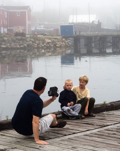 Man photographing two boys sitting on edge of dock with water and dock in background on foggy day